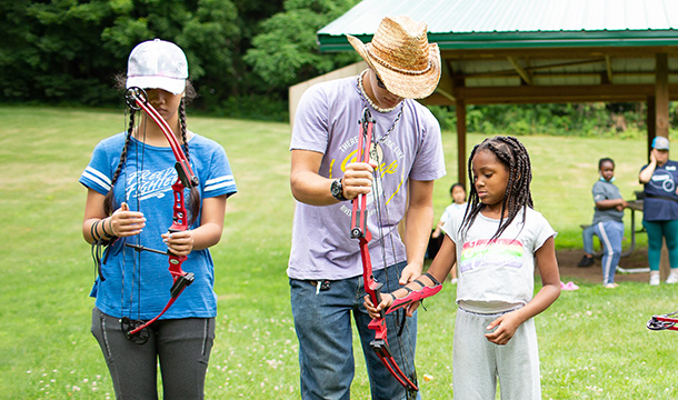 Volunteer in straw hat teaches campers archery 