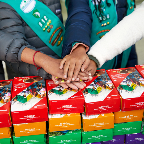 Girls showing teamwork with hands in a huddle on top of Girl Scout Cookie Boxes