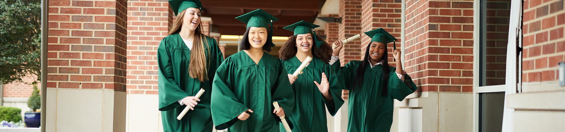  Girl Scouts in green graduation cap and gowns holding paper scrolls and smiling outside campus 