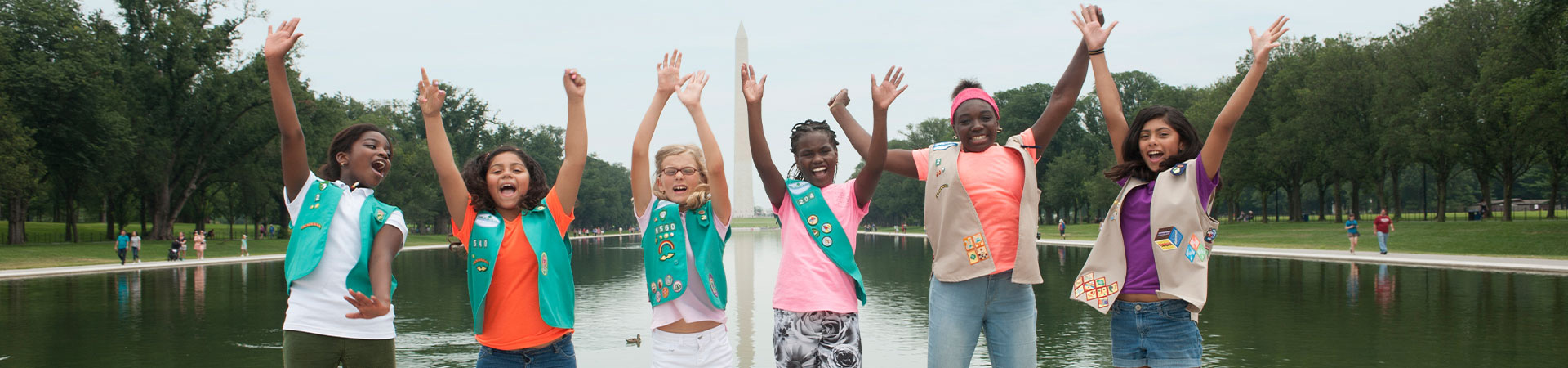  Girl Scouts in uniforms jumping with arms up in front of water 